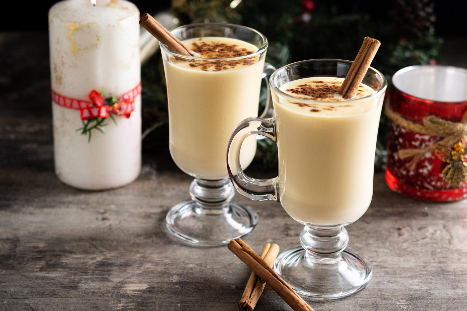 Two glass mugs of creamy eggnog topped with ground nutmeg and garnished with cinnamon sticks, set on a rustic wooden table with holiday candles and greenery in the background