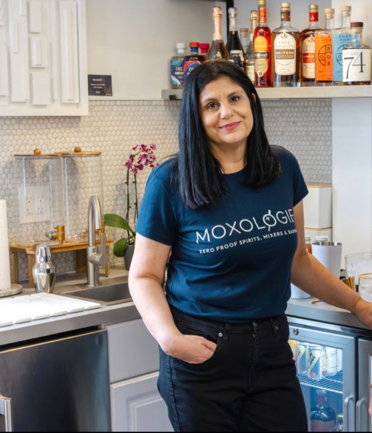 Person wearing a blue 'Moxologie' t-shirt in a kitchen setting with bottles on the counter.